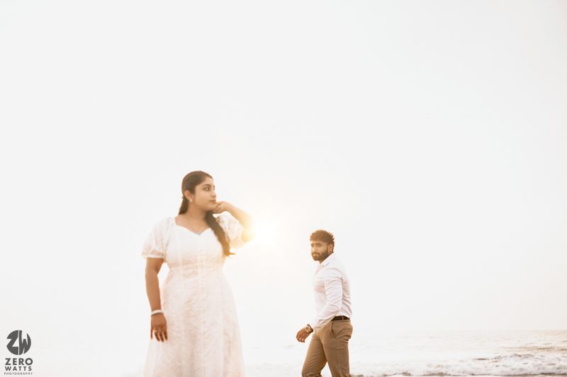 A couple stands together at the beach during sunset wearing white outfits against a bright background.