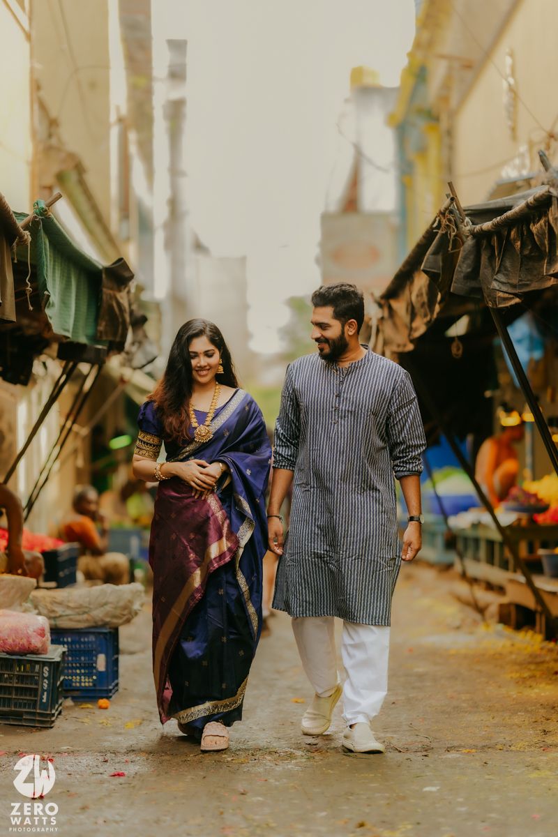 A couple walks hand in hand through a bustling street market with vendors and stalls visible in the background.