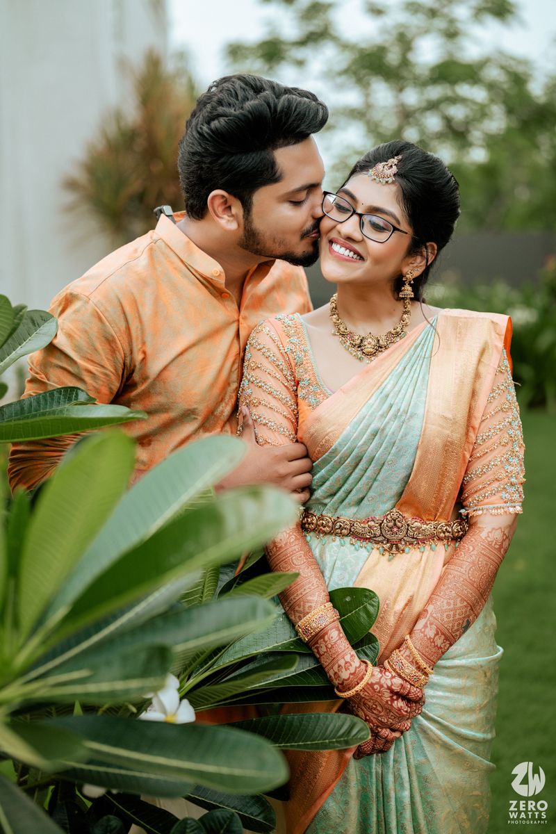 Traditional Indian couple shares intimate moment in orange and mint outfits amidst tropical garden setting.