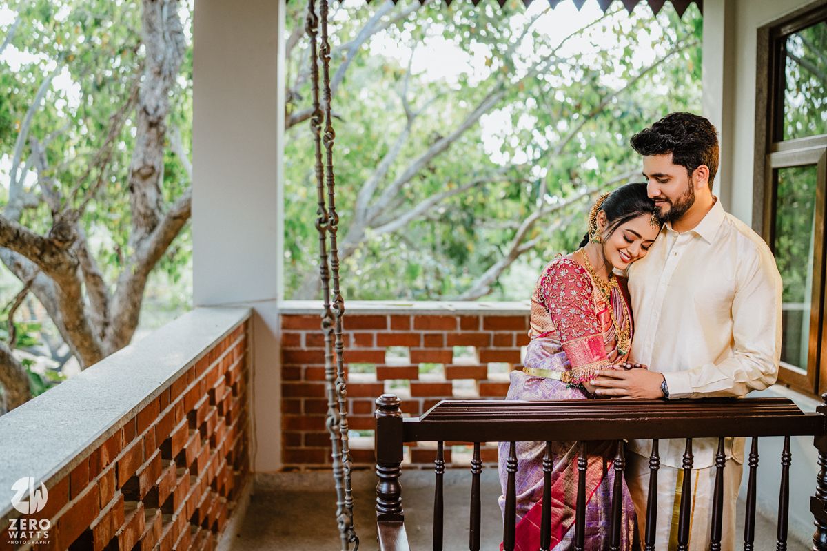 A romantic couple sharing an intimate moment on their balcony with brick railings and lush greenery in the background.