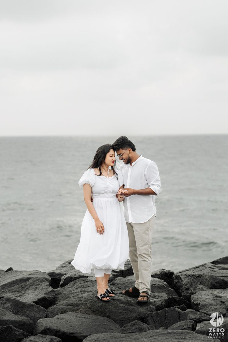 A romantic couple stands on dark coastal rocks against a moody gray ocean backdrop.