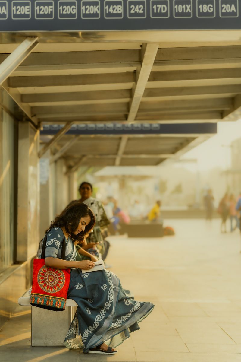 Person in traditional dress sits at a subway station platform with a decorative handbag in soft sunlit atmosphere.