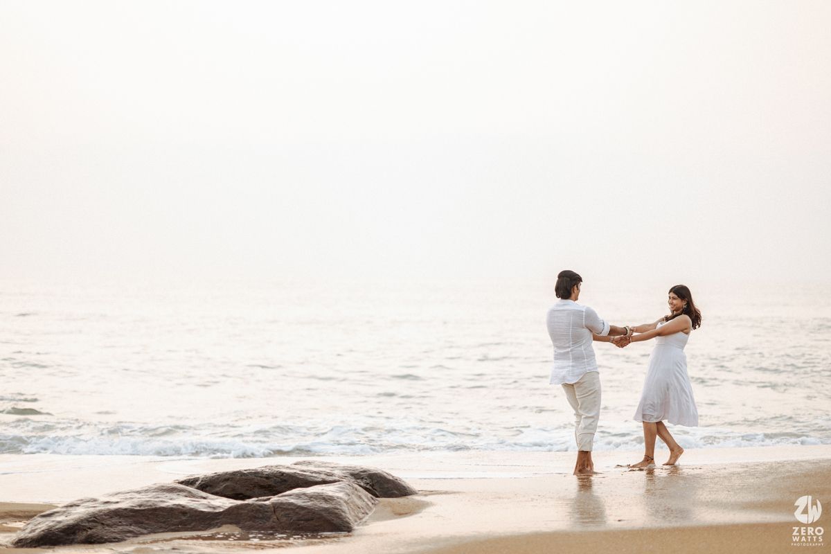 Two people in white clothing dancing together on a beach near rocks at dusk.