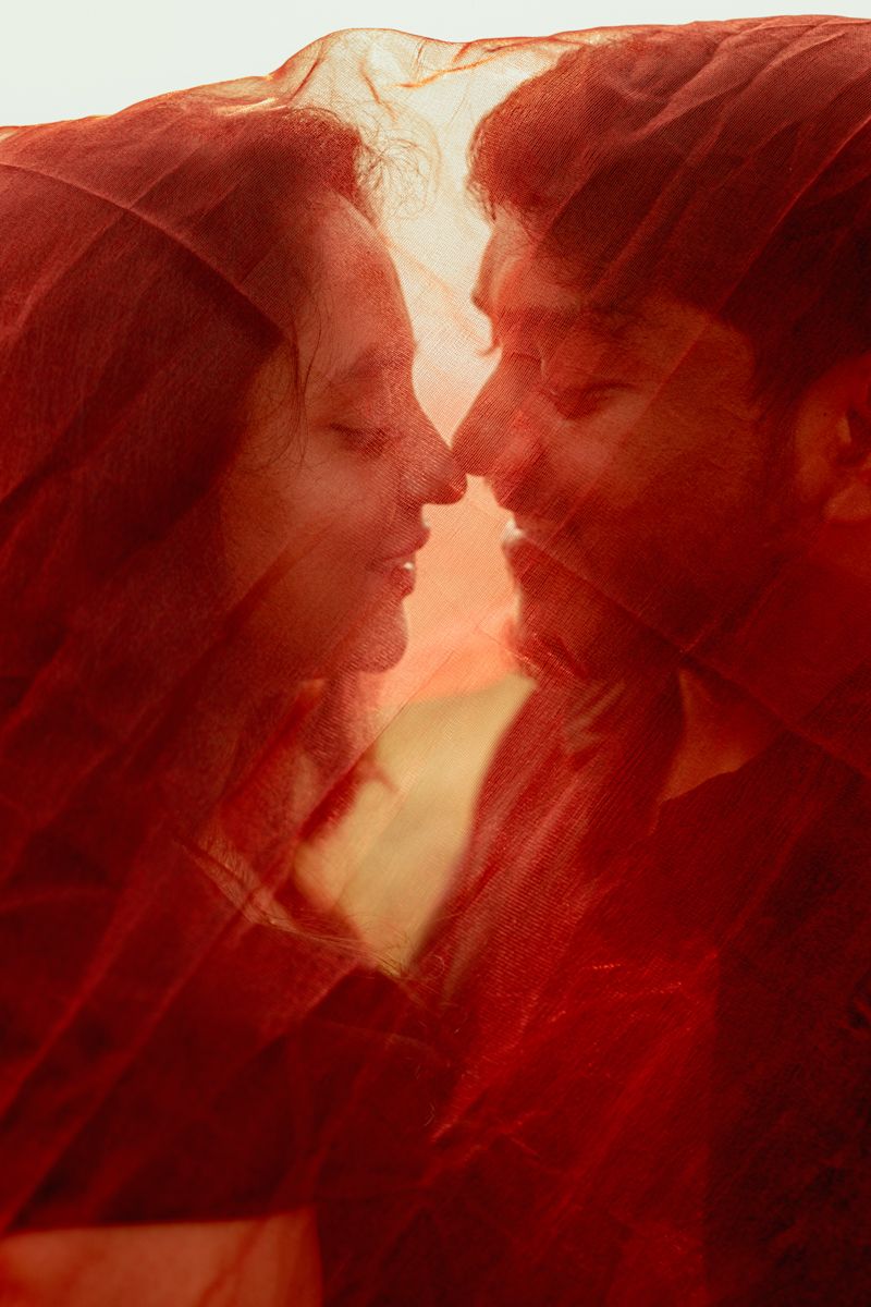 Romantic couple sharing an intimate moment through red sheer fabric creating a dreamy atmosphere.