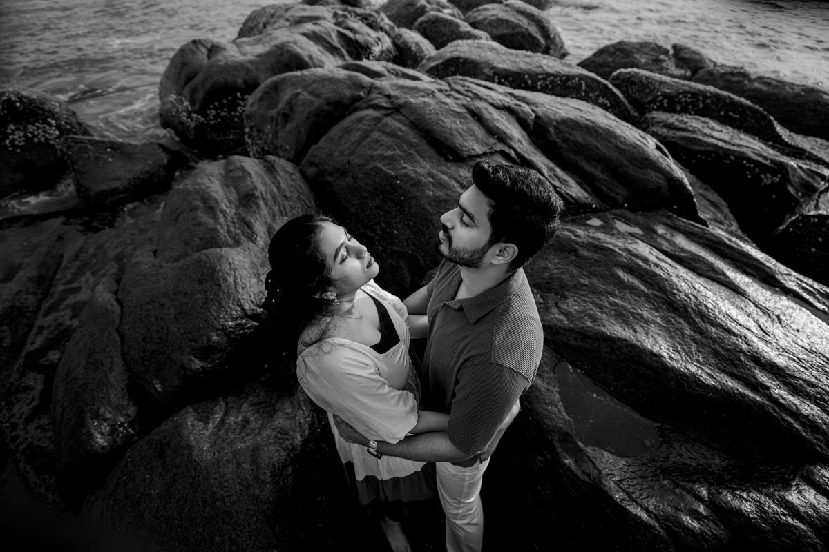 A romantic black and white photo of a couple embracing on rocky coastal terrain.