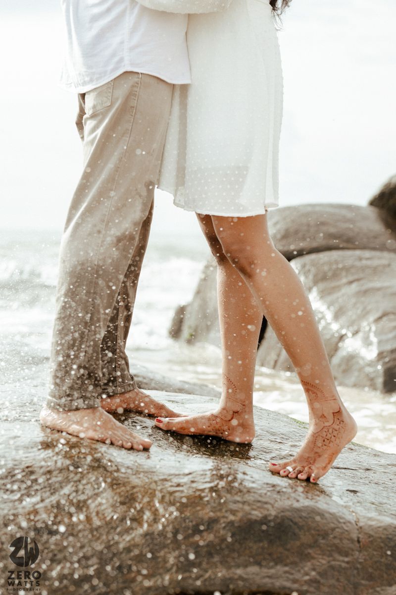 Close-up of bare feet standing on wet rocks by the ocean with water splashing around them.