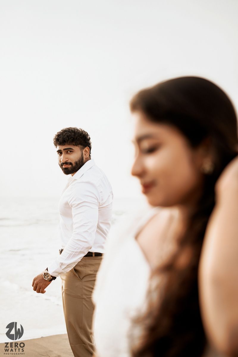 A romantic beach photoshoot featuring two people in light colored outfits against an overcast sky.