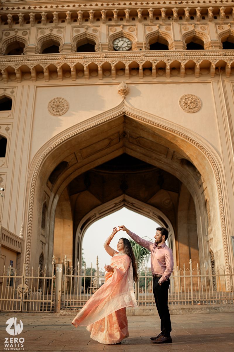 A couple dances beneath an ornate archway of historical Indian architecture, with the woman wearing a flowing peach gown.
