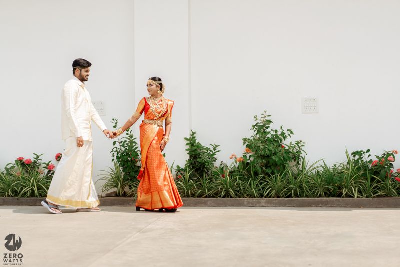 A couple in traditional Indian wedding attire holds hands against a white wall with tropical greenery and flowers.