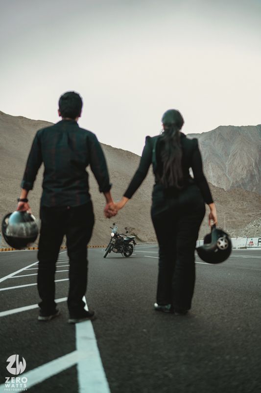 Two riders in black holding helmets walk together on a mountain road with their motorcycle in background.