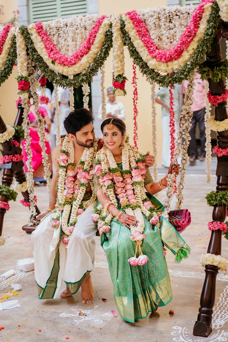 Couple on decorated wedding swing adorned with pink and white flower garlands during traditional South Indian ceremony.