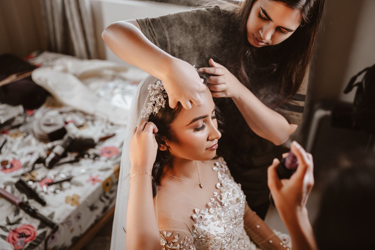 A stylist prepares a bride wearing an elegant lace gown for her special day in a dimly lit room.