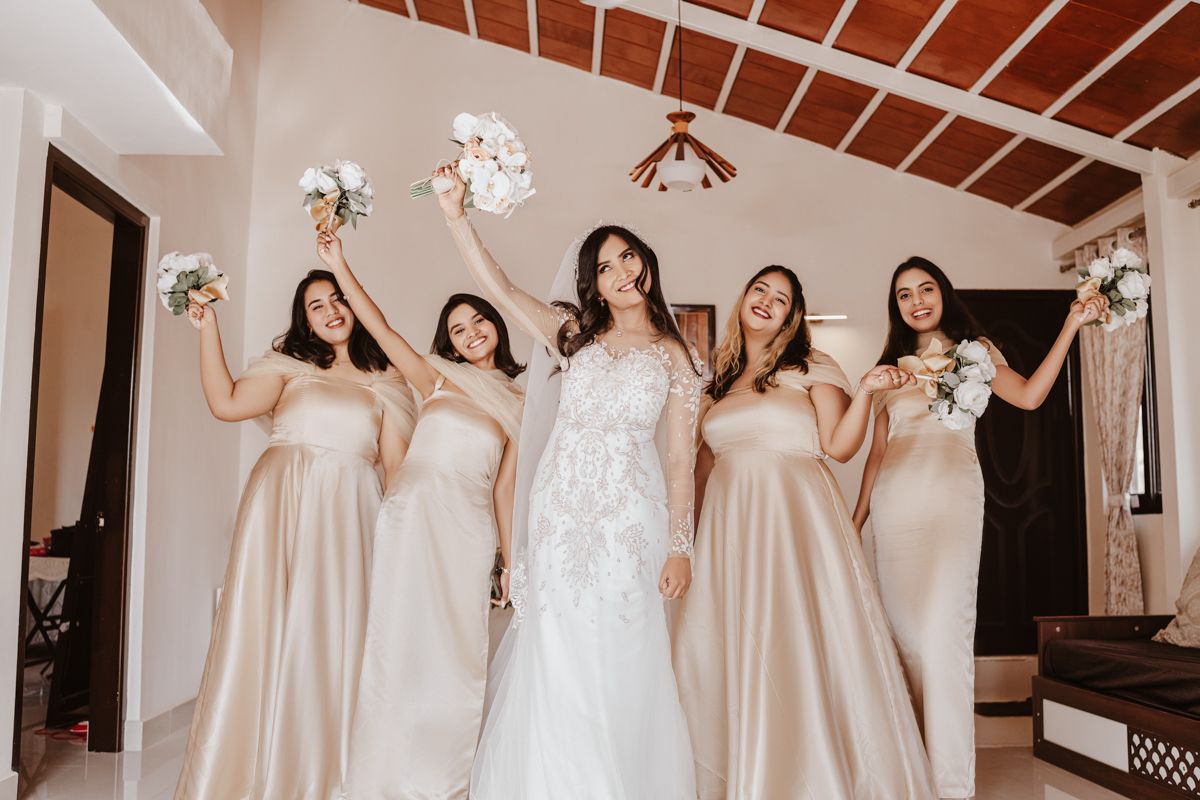A wedding party poses together in elegant white and champagne gowns while holding bouquets in a sunlit room.