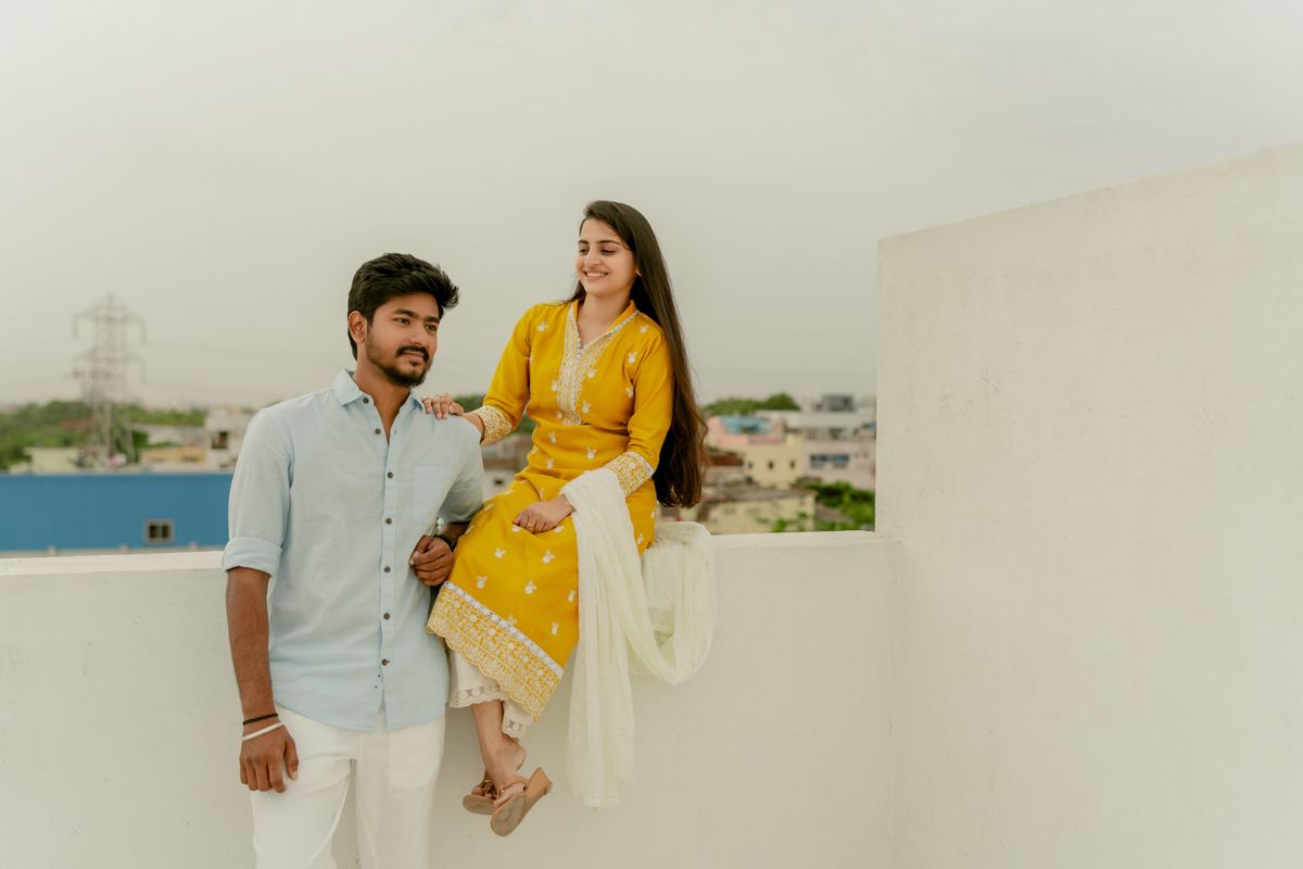 A couple in coordinated casual outfits pose playfully against a white wall with a city view in the background.