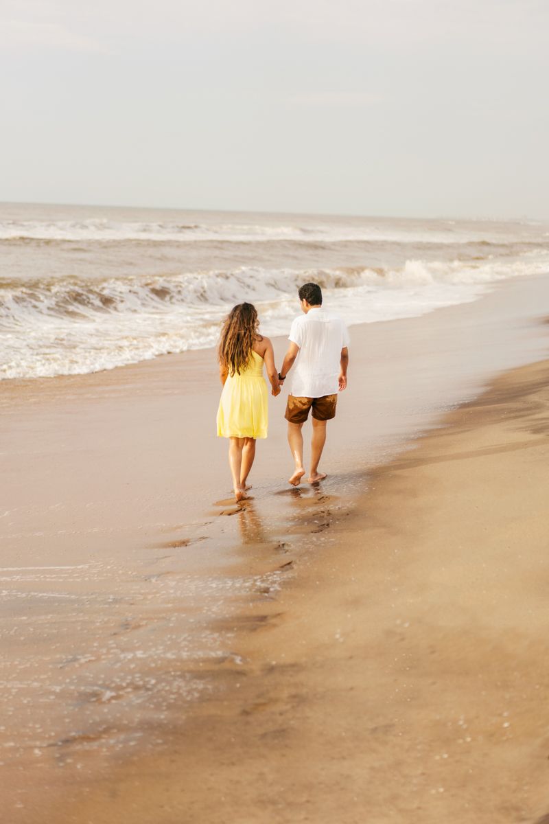 A couple walks together along a sandy beach with crashing waves as the woman wears a yellow dress and man in white shirt.