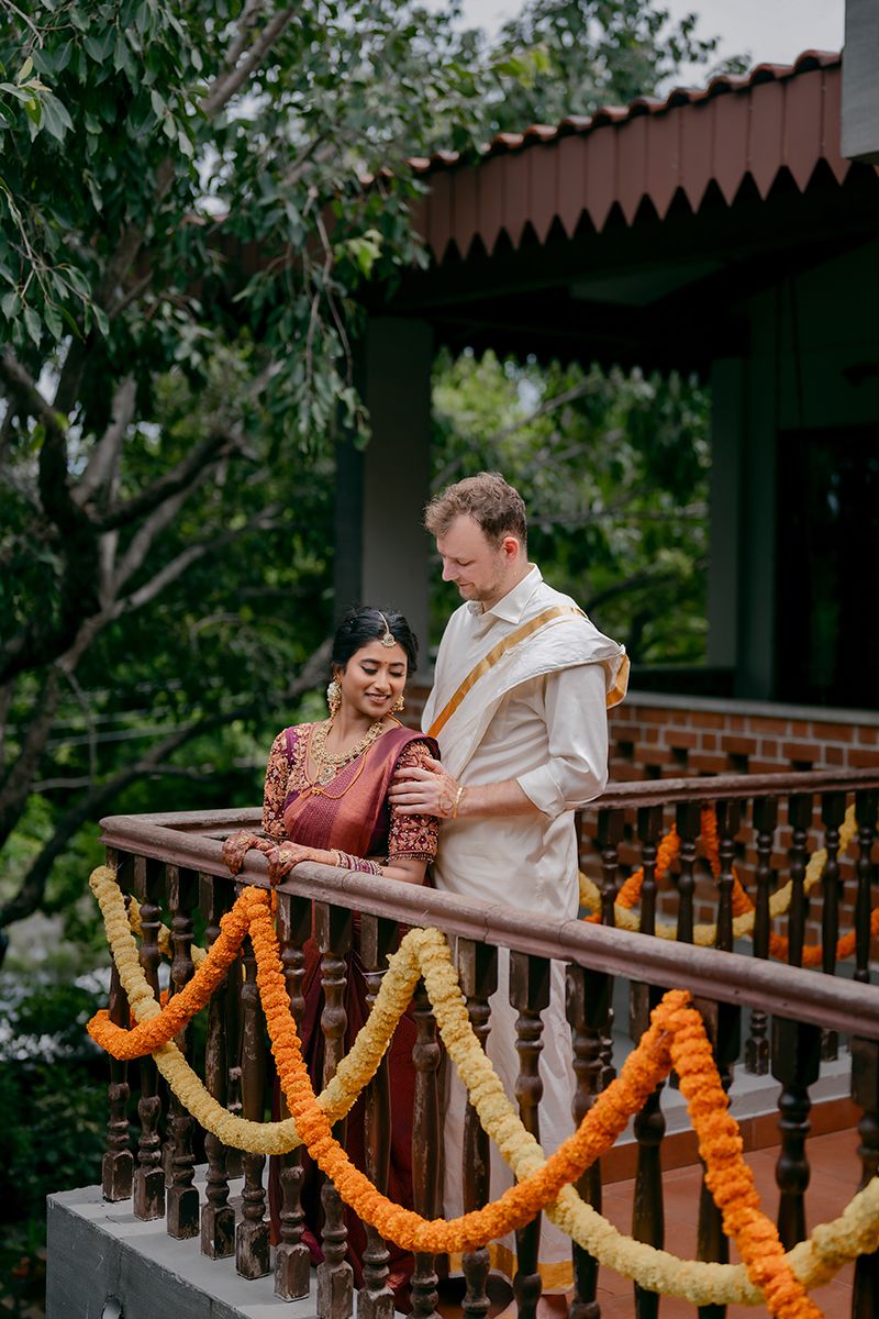 Couple in traditional wedding attire standing on a decorated veranda with marigold garlands during an Indian fusion ceremony.