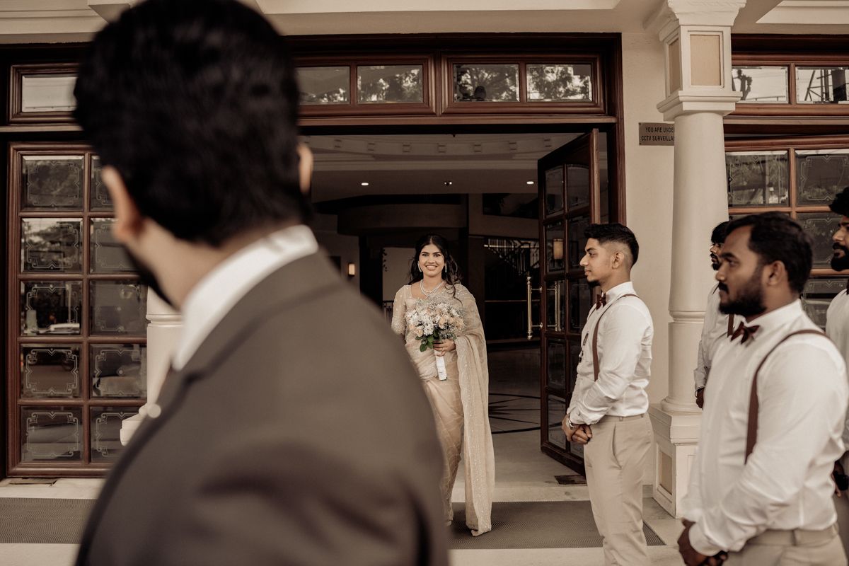 Elegant wedding ceremony scene with figures in neutral tones standing before ornate brown wooden doors at traditional venue.