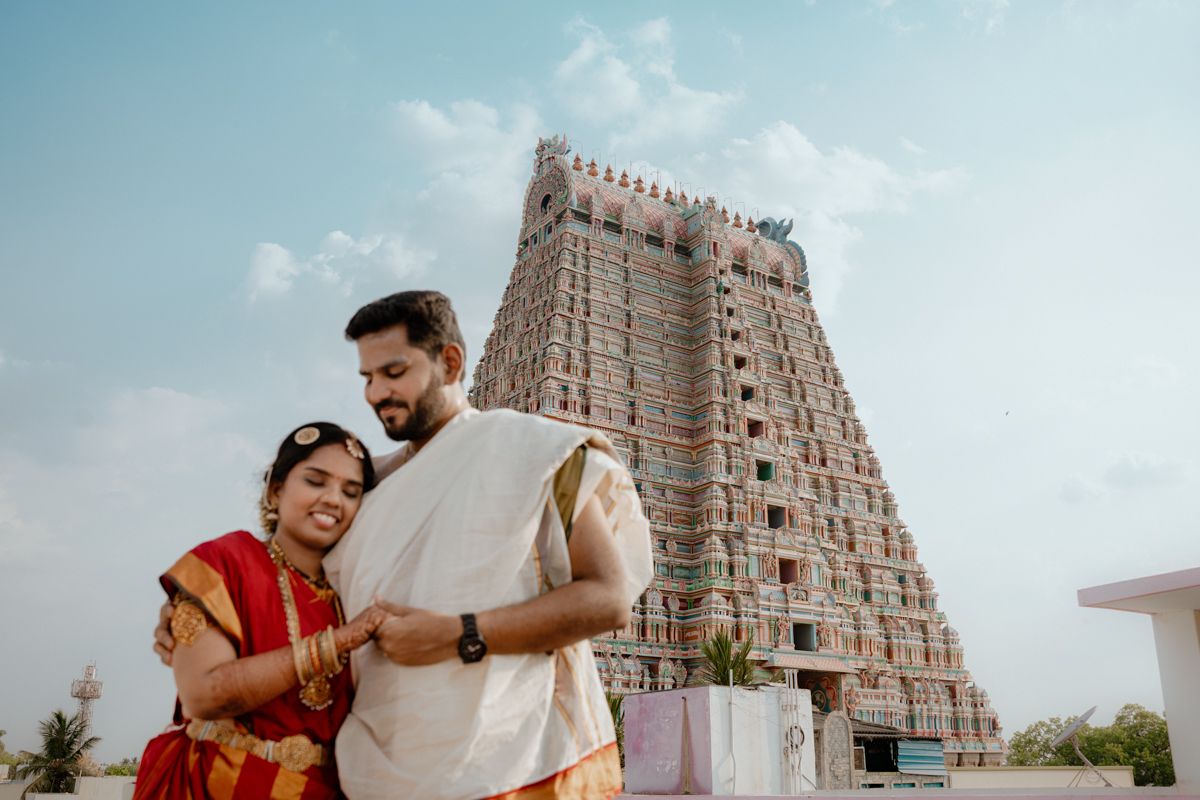 A couple in traditional and casual attire stand together in front of a towering ancient temple gopuram in South India.