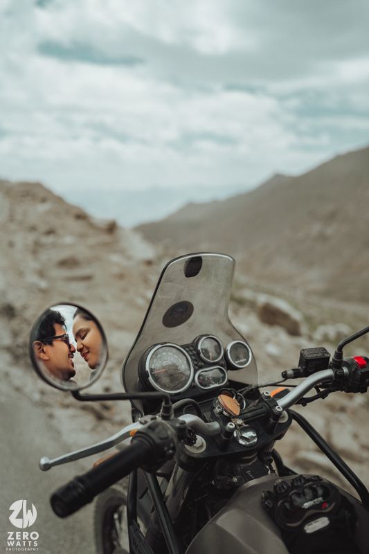 A motorcycle's rearview mirror reflects a mountain landscape under cloudy skies with handlebar controls visible.