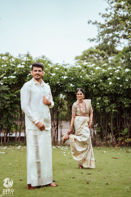 Traditional South Indian couple in white and gold attire pose in a garden setting with blooming flowers.