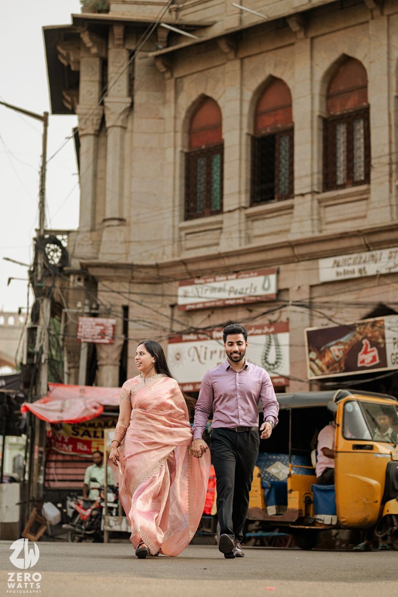 Romantic couple walking through busy Indian street with colonial architecture and auto rickshaw in background.