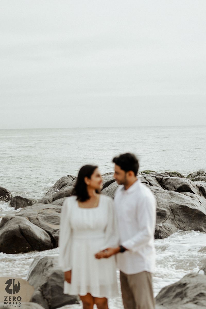 A couple in white clothing stands intimately on rocky coastal beach with misty grey ocean backdrop at dusk.
