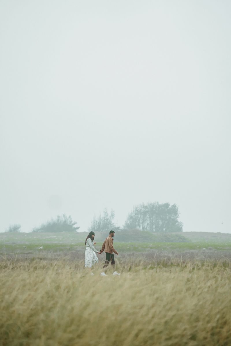 A couple walks together through a misty field of tall grass on an overcast day.