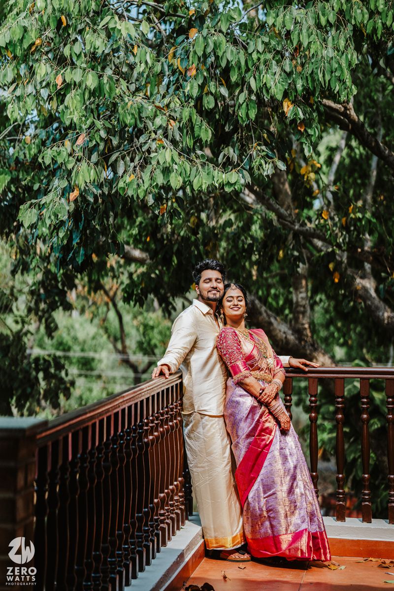 Newlyweds in traditional attire pose on wooden bridge surrounded by lush trees.