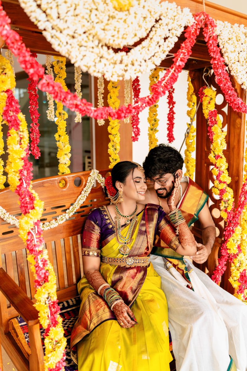 Couple sharing a candid moment on a wooden swing adorned with yellow and red flower garlands at a traditional event.