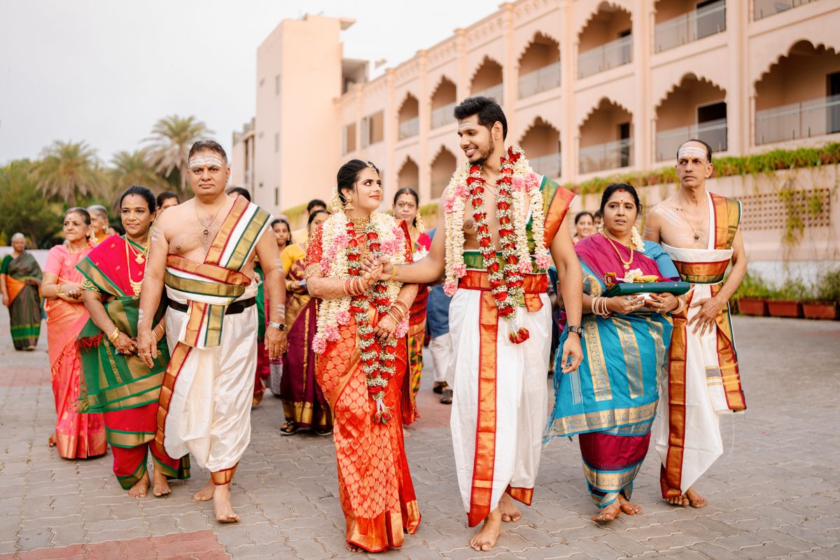 A traditional South Indian wedding procession with people wearing colorful saris and garlands at a temple-like venue.