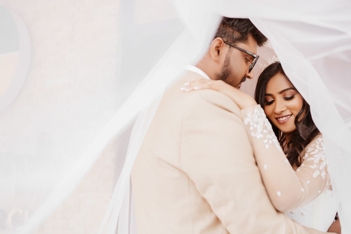 A romantic wedding portrait under flowing white veil showing tender embrace between newlyweds.