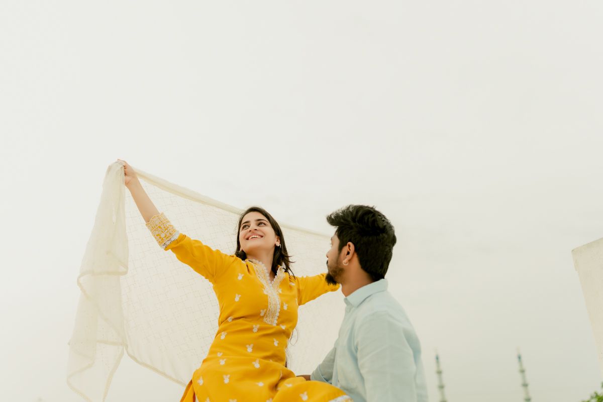 A couple in yellow and white outfits share a playful moment on a rooftop against an overcast sky.