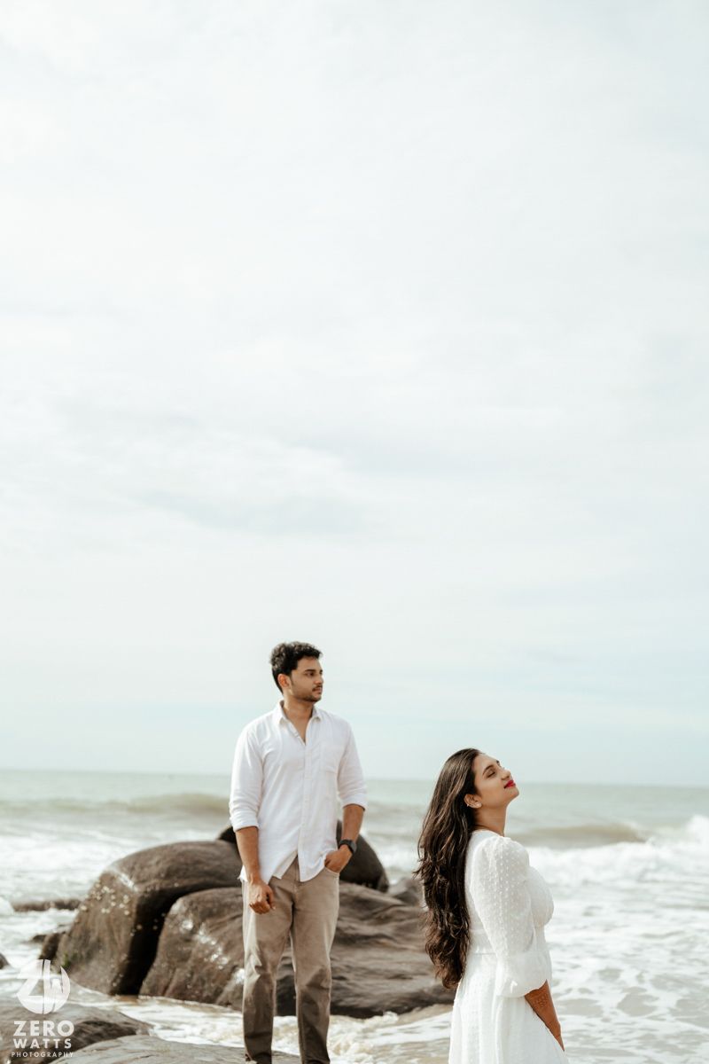 A couple in white and beige outfits poses together on rocky beach with ocean waves in the background.