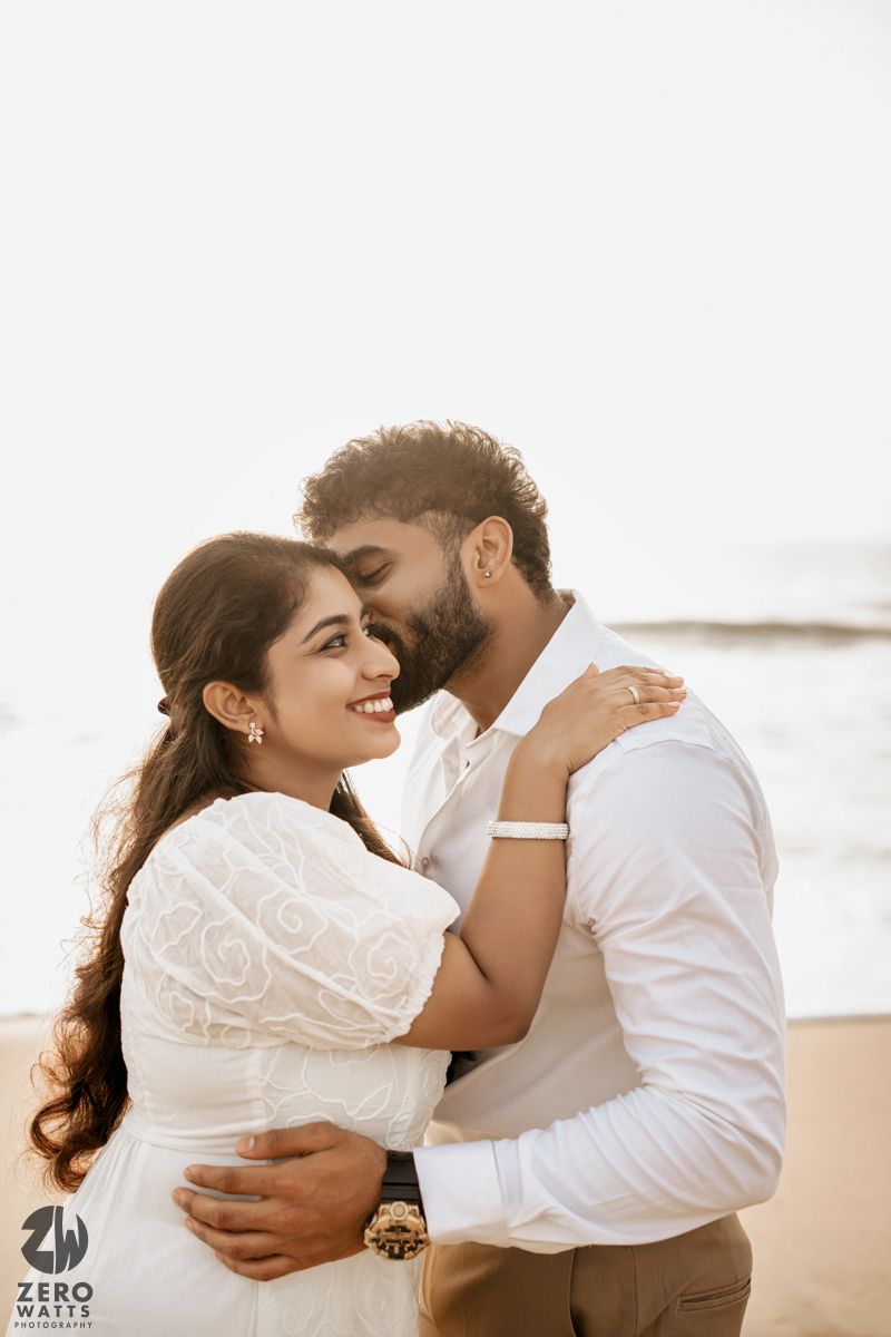 A romantic couple embraces on a beach at sunset, with the woman wearing a white lace dress.