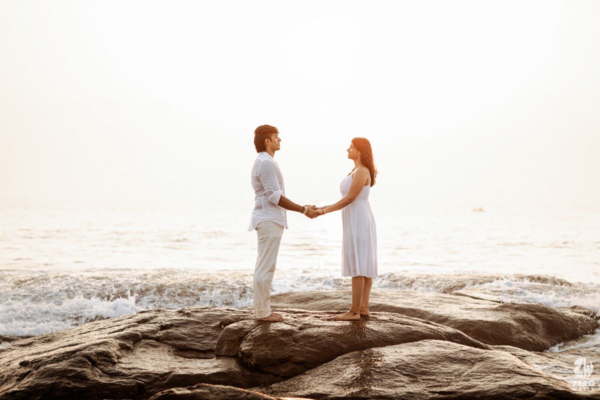 Romantic couple holding hands while standing on rocky beach during sunset by the ocean.