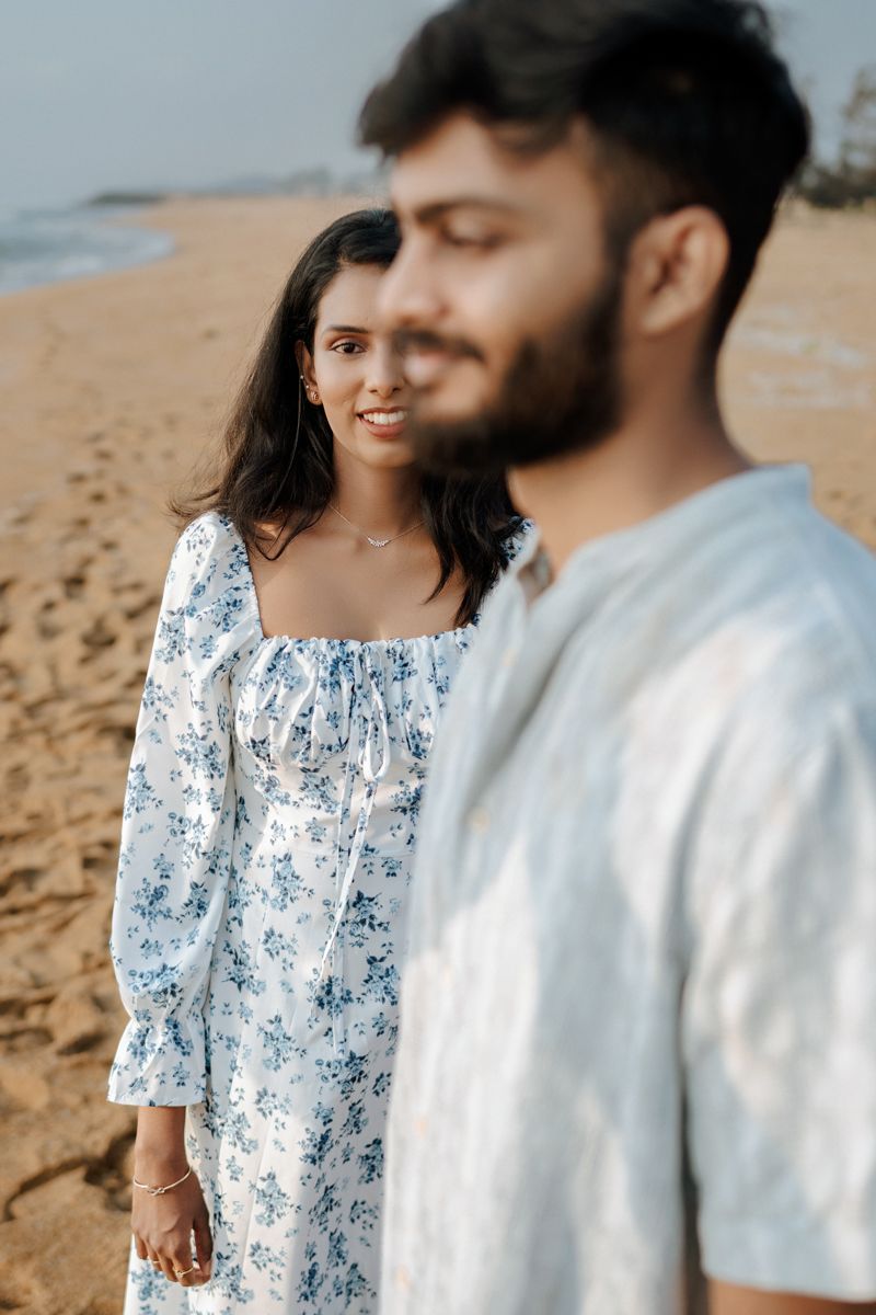 A romantic couple stands together on a sandy beach, with her wearing a white floral dress and him in casual attire.