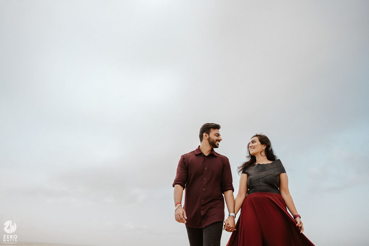 A couple in brown and burgundy outfits hold hands and gaze at each other against a cloudy sky.