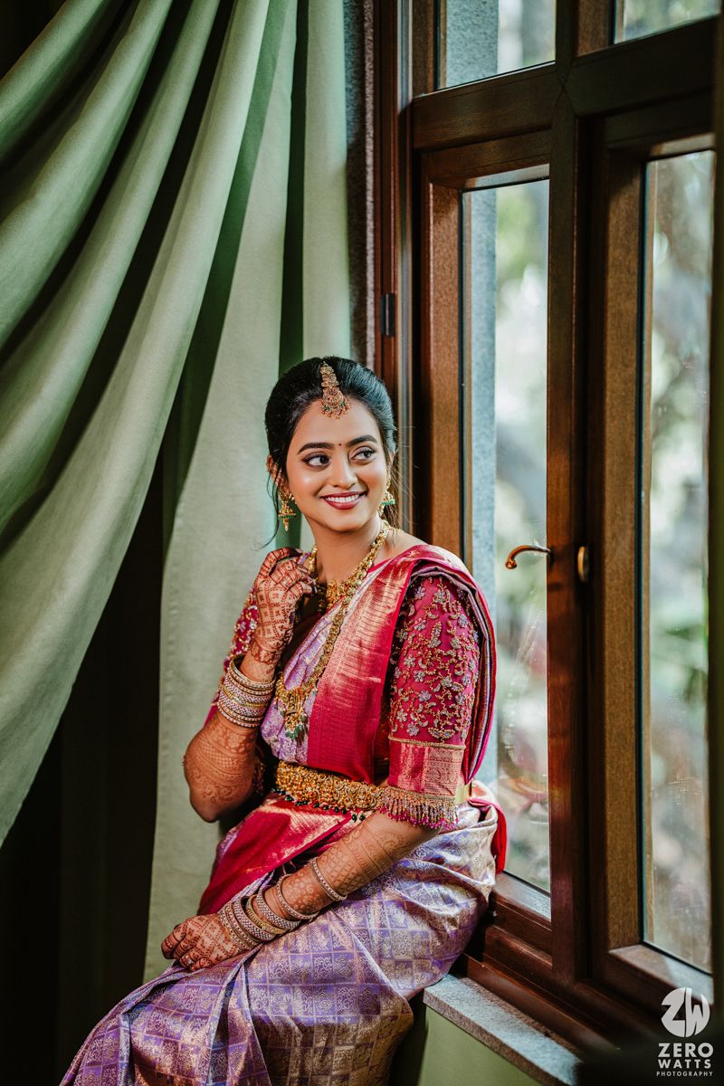 Candid moment of traditional bride in silk saree and henna sitting by window with natural light.