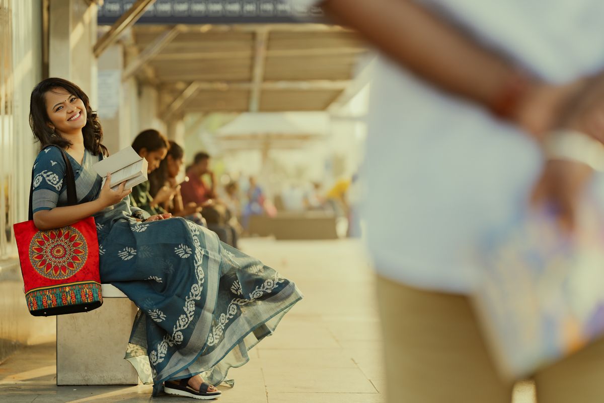 Person in blue dress sits on bench with traditional style bag while others rest in background at station platform.