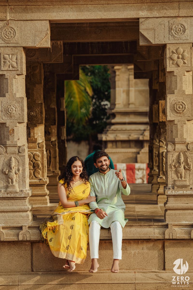 A couple in yellow and mint outfits sit together on stone steps of an ancient temple corridor.