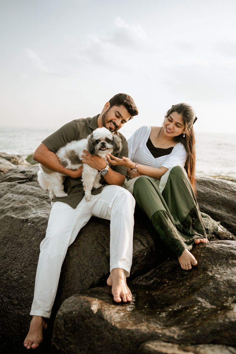 Couple sits on coastal rocks with their small white dog against an ocean backdrop.