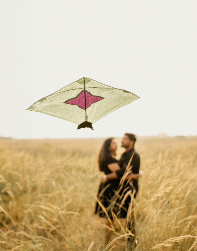 A simple kite with geometric design floats above a couple embracing in a golden wheat field at sunset.