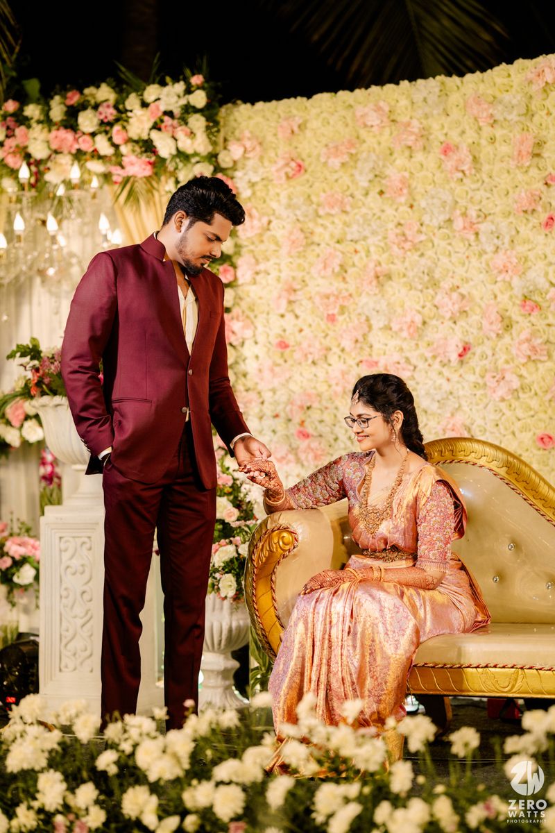 An elegant couple at their engagement celebration with a maroon suit and peach gown against a floral wall backdrop.