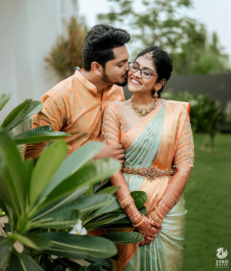 A couple in matching peach colored traditional outfits share a tender moment in a garden setting with tropical plants.
