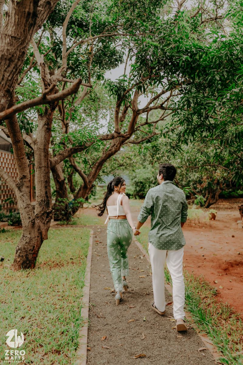 A couple holds hands while walking along a dirt path under green trees in a romantic outdoor setting.