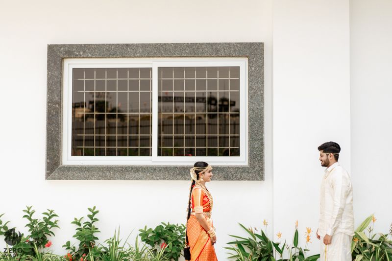 Two people in traditional Indian wedding attire stand against a white wall with decorative window and greenery.
