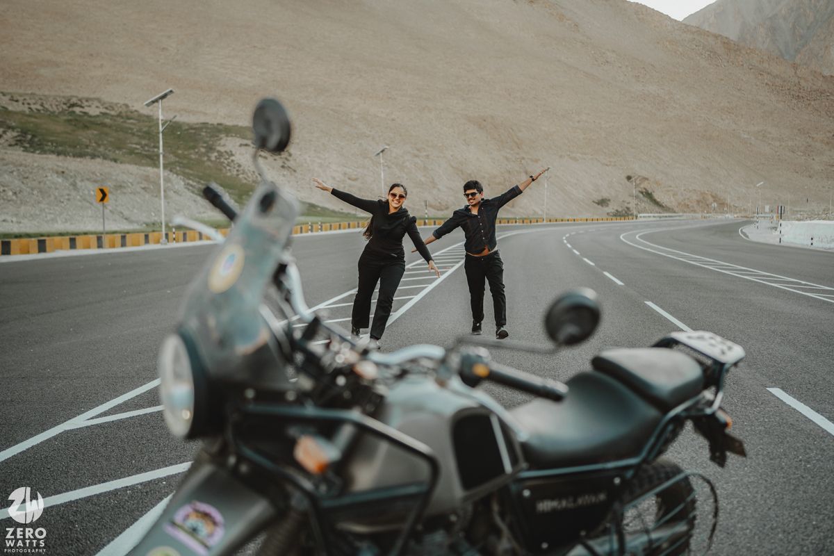 Two people standing with arms raised on empty mountain highway near parked motorcycle.