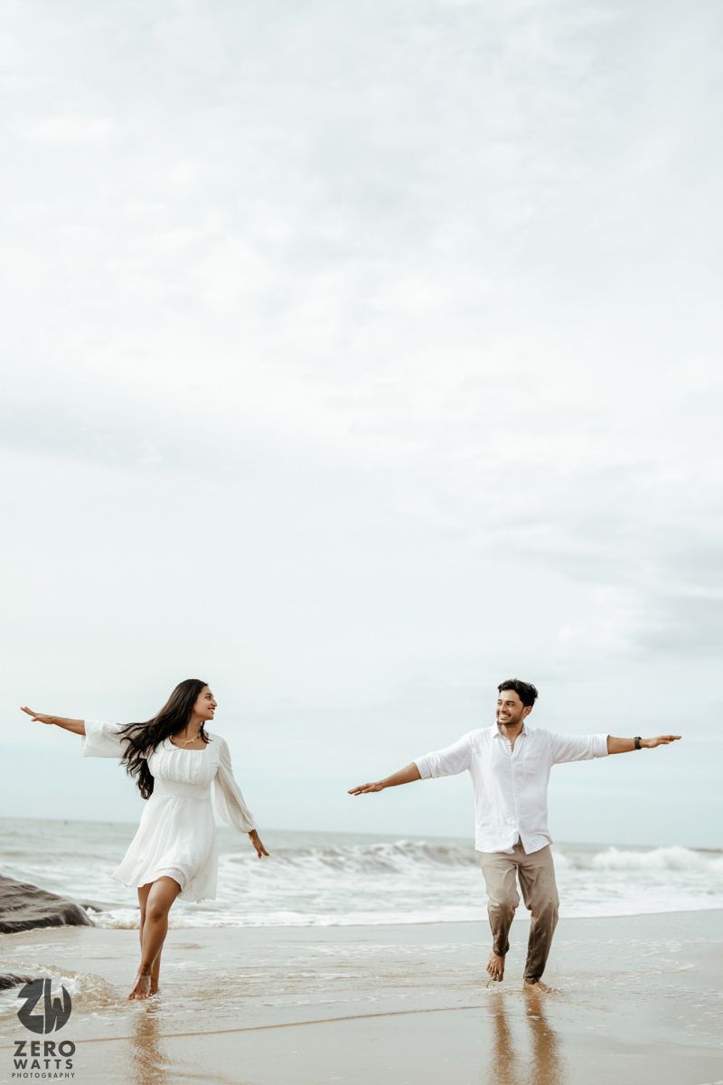 A couple in white outfits dances joyfully on a beach with arms outstretched against an ocean backdrop.