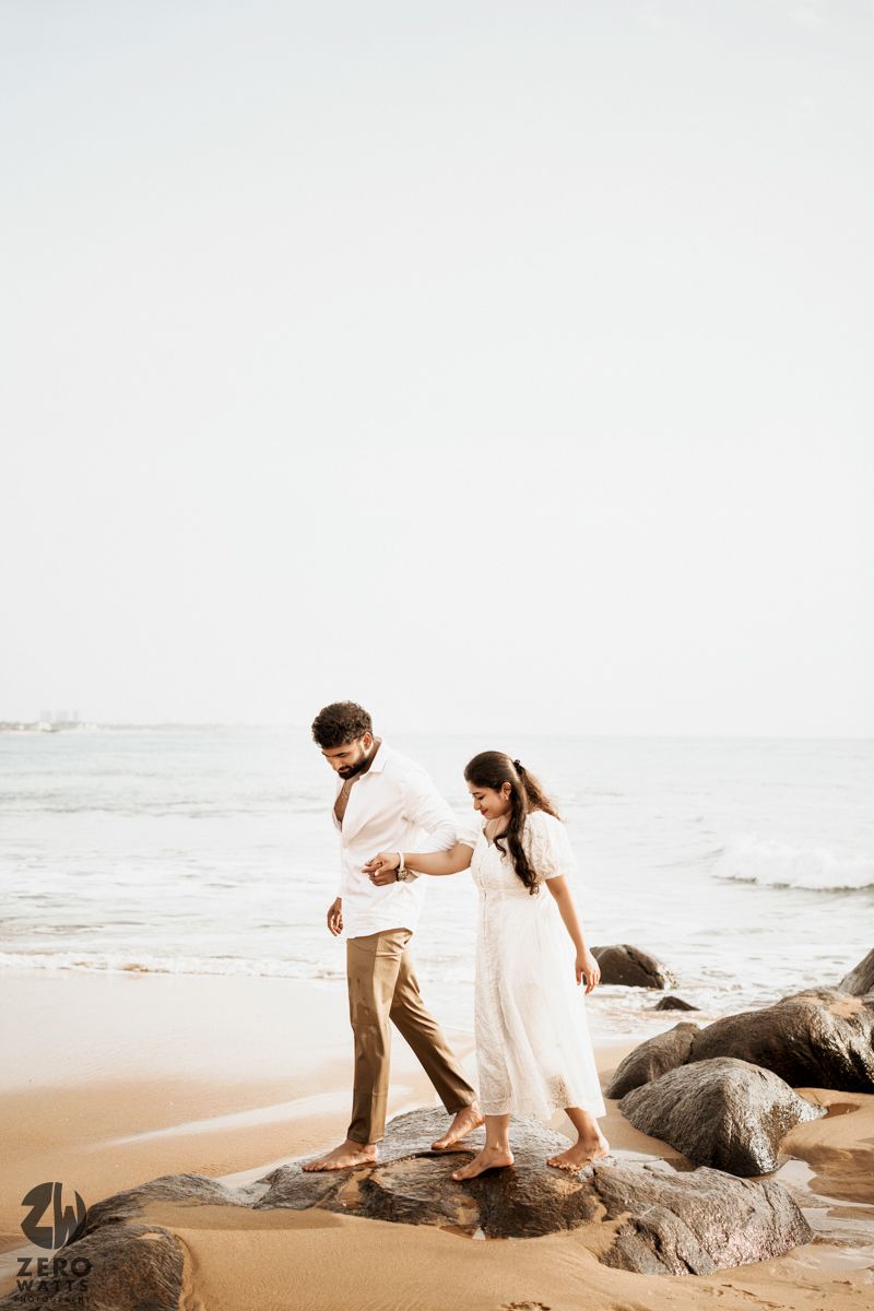 Couple walking hand in hand along rocky beach during photoshoot.