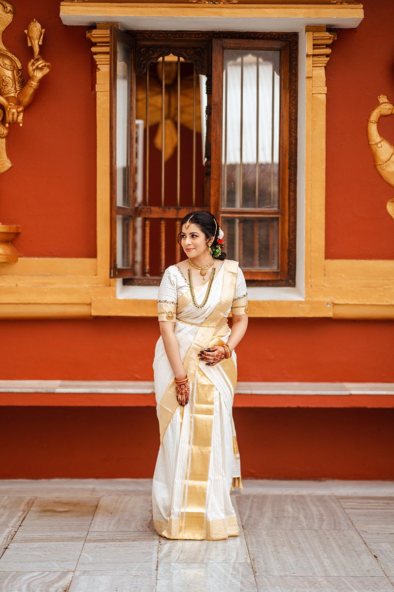 Traditional South Indian bride in cream and gold kasavu saree stands against terracotta wall with vintage window.